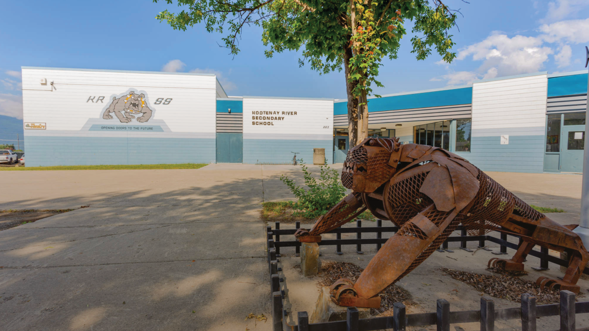 A flat-roofed blue and white school building against a blue sky with a leafy green tree and a rust-coloured sculpture of a bulldog in the foreground