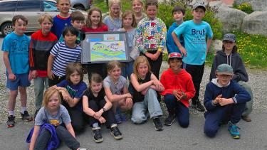 A group of elementary school students pose in front of a display board in a park