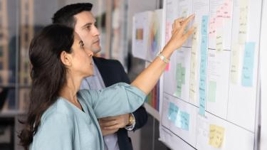 A woman with long dark hair and a man with short dark hair plan at a whiteboard using stickies.