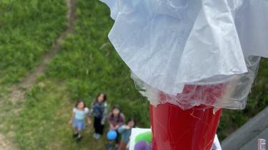 A close up of a person's hand holding a styrofoam square with a red cylinder on top attached to a balloon on top of a mechanical lift.