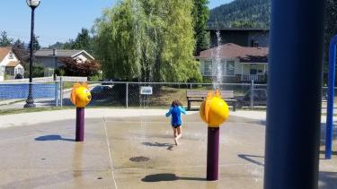 A child in a blue shirt and black shorts cavorts in a spray park in the sunshine